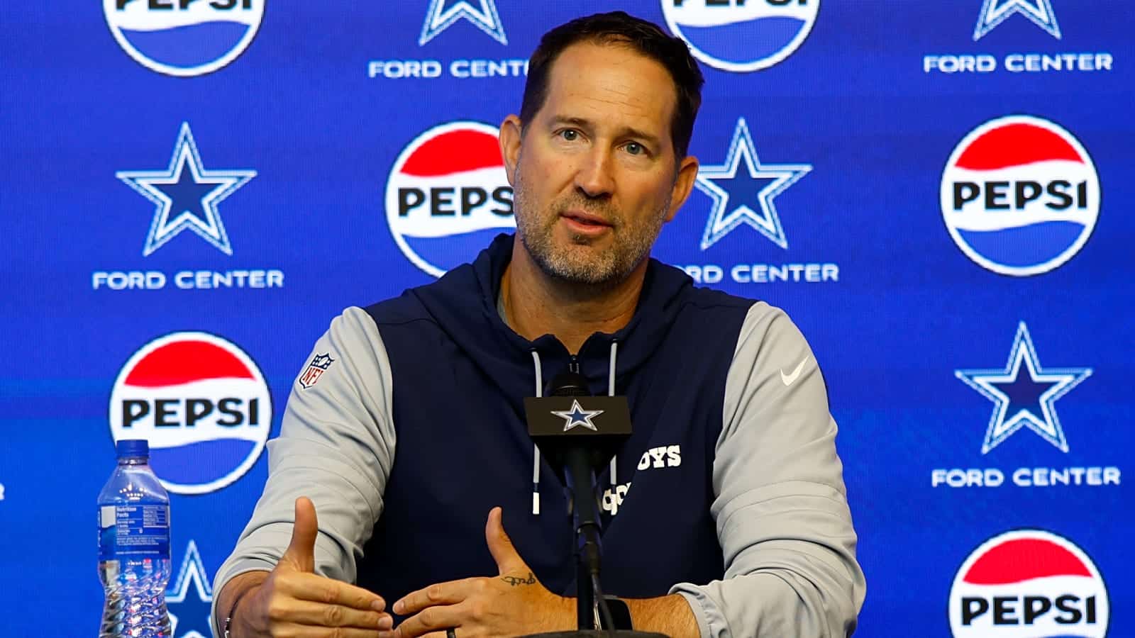 Dallas Cowboys head coach speaking at a press conference at Ford Center, with Pepsi and Cowboys branding in the background. Focus on NFL coaching staff, team updates, and inside the star sports coverage.