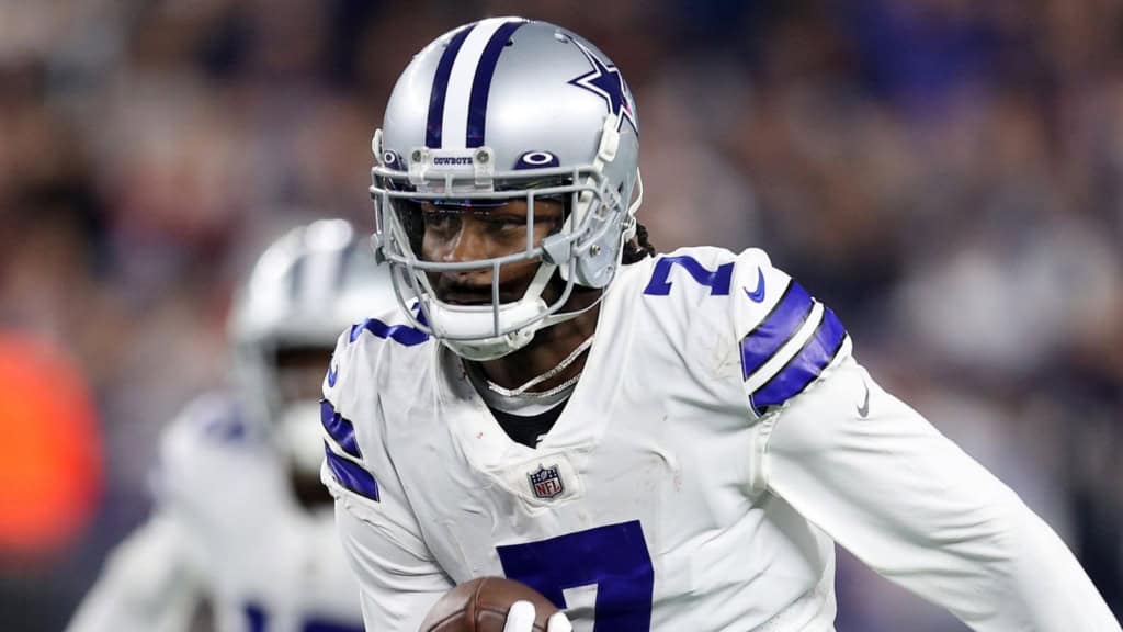 Dallas Cowboys football player in action during a game, wearing team uniform and helmet, holding football, focused on play, inside the stadium with blurred crowd background.