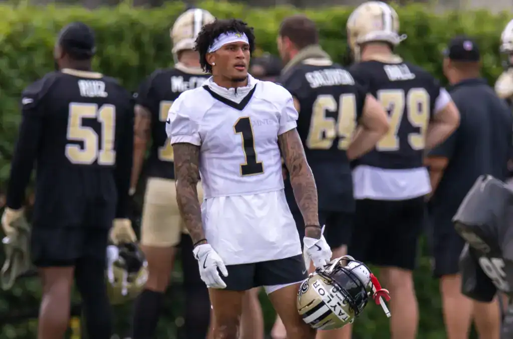 Young football player wearing a white jersey with the number 1 during practice, with teammates and coaches in black and gold uniforms in the background, holding helmets, outdoors during the day.