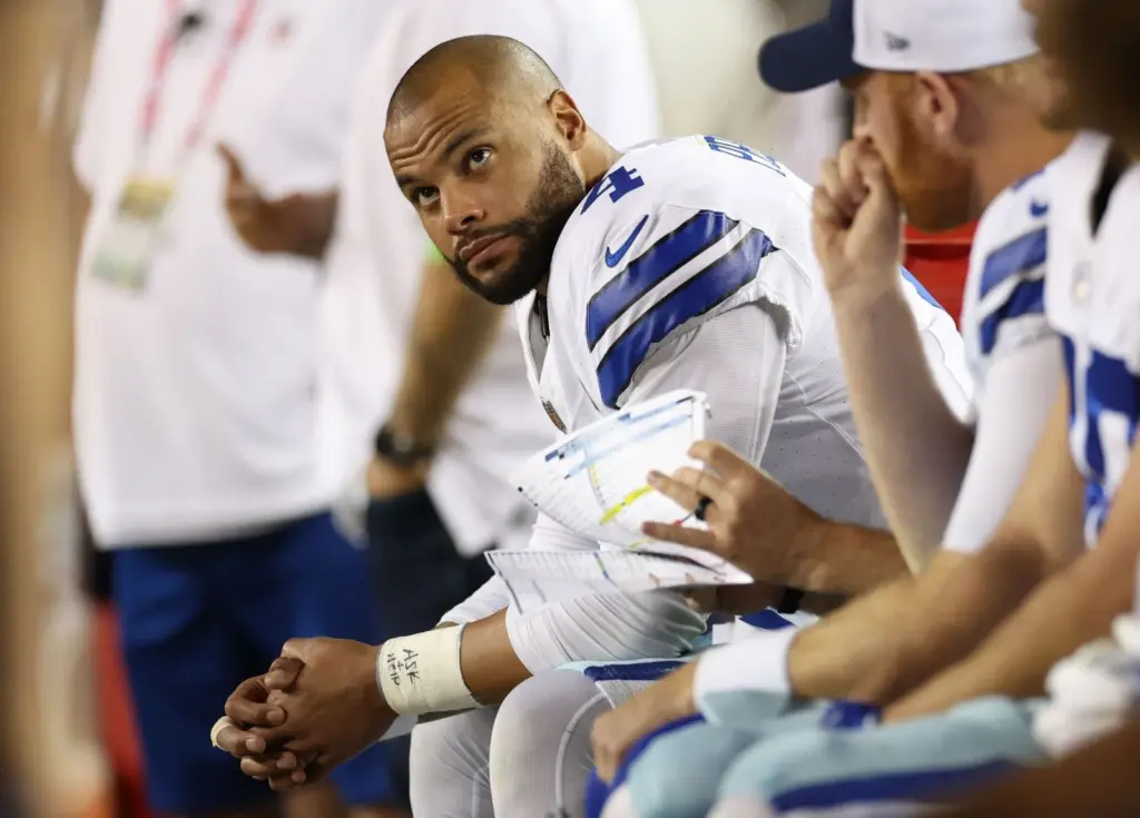 A Cowboys football player during a game meeting with coaches on the sideline, wearing a white jersey with blue and gray accents, holding play sheets, in a focus moment.