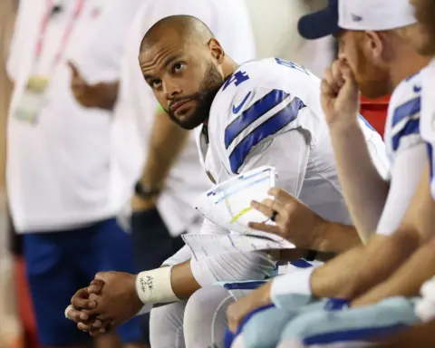 A Cowboys football player during a game meeting with coaches on the sideline, wearing a white jersey with blue and gray accents, holding play sheets, in a focus moment.