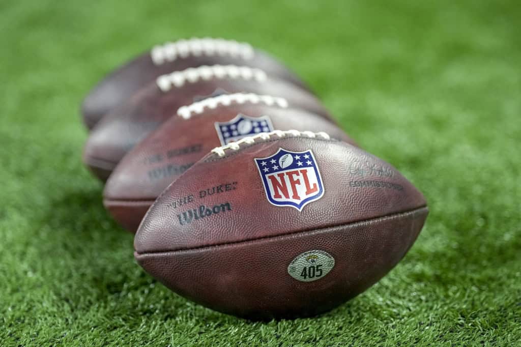 Close-up of four NFL footballs lined up on a green grass field, showcasing the official NFL logo, brand markings, and textured leather surface perfect for football game action pictures.