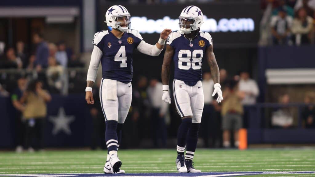 An image of two Dallas Cowboys football players in navy blue and white uniforms during a game, with one player gesturing towards the other as they celebrate a play.