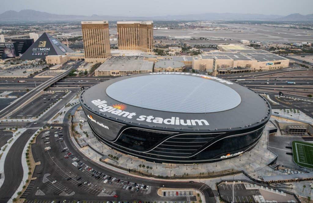 Sleek aerial view of Allegiant Stadium in Las Vegas, home to the Las Vegas Raiders NFL team, featuring its modern architecture, surrounding parking lots, and nearby city skyline.