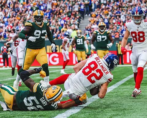 A Dallas Cowboys player with jersey number 88 is reaching for the end zone as he is tackled by a Green Bay Packers defender, number 55, during an exciting NFL game at Lambeau Field with a packed crowd cheering in the background.