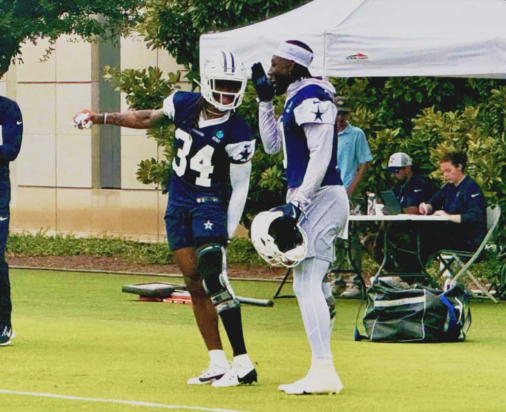 An image of two Dallas Cowboys football players in practice gear, one with a helmet and the other conversing, on a practice field with team staff and equipment in the background.