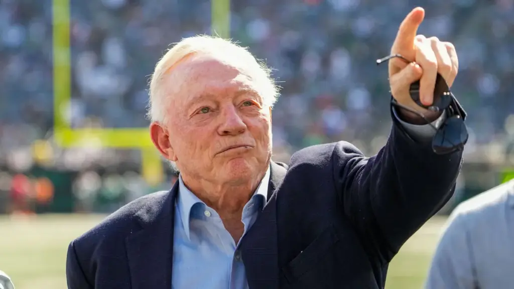 Senior man pointing with expression at an outdoor football event, wearing a suit jacket and blue shirt, with football field in background.
