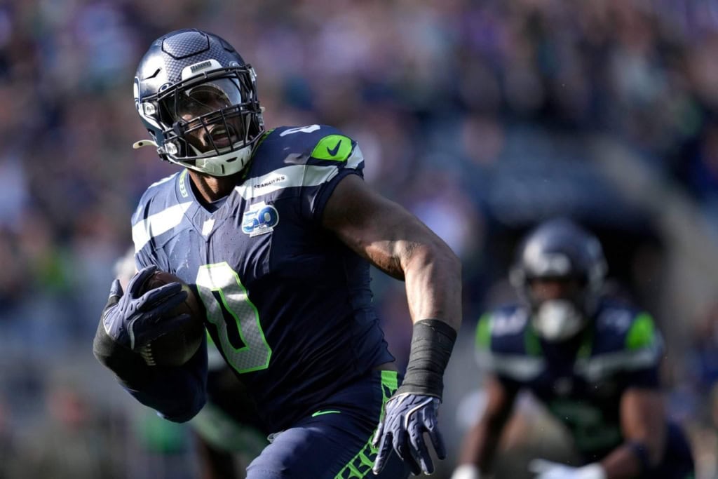 Seahawks football player running with ball during NFL game, team in blue and green uniform, stadium crowd in background.