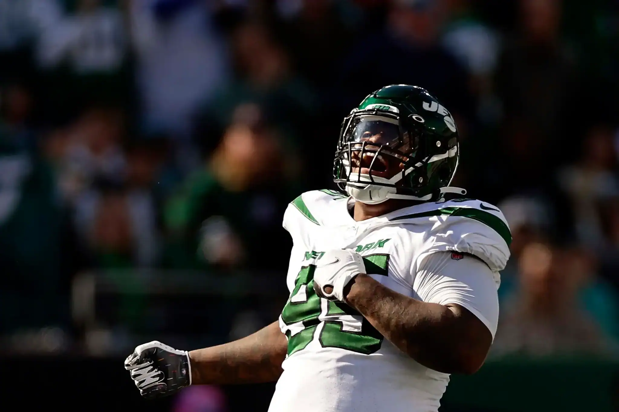 A football player from the Dallas Cowboys celebrating during a game, wearing a white jersey with green accents, helmet, and gloves. The background is blurred with fans in the stands.