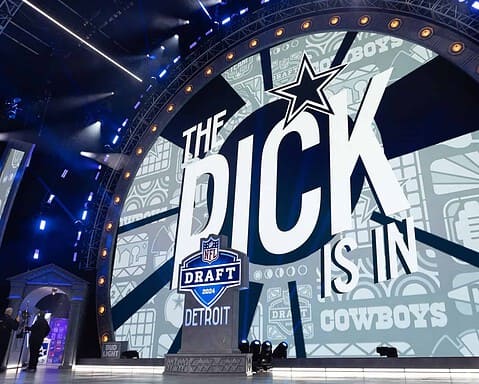 Draft NFL 2024 NFL Draft event stage with prominent Dallas Cowboys branding, large digital screen displaying "The Pick is In," surrounded by professional lighting and event setup.