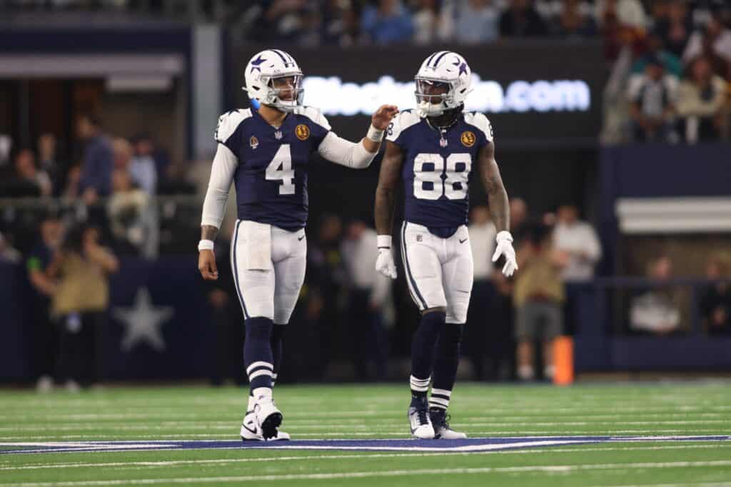 Dallas Cowboys football players celebrating on the field during a game, wearing navy blue and white uniforms, with a blurred crowd in the background.