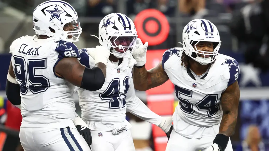 Celebrating Dallas Cowboys football players on the field wearing white jerseys and helmets with star logos, showcasing team camaraderie, during an NFL game.