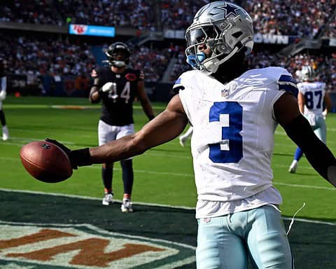 A professional football player from the Dallas Cowboys holding a football on the field during a game. The player is wearing a white jersey with the number 3, helmet, and black gloves, with other players and fans visible in the background.