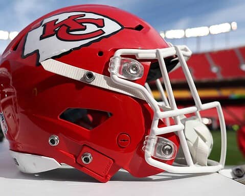 A close-up image of a red Kansas City Chiefs NFL helmet featuring the team logo and white facemask, displayed on a table at a football stadium during daytime.