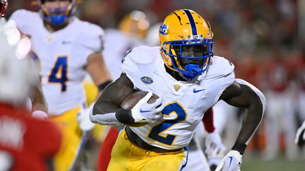 A college football player from the University of Georgia (UGA) in yellow and white uniform carries the football during a game, showcasing athleticism and team spirit, with other players and the stadium background.