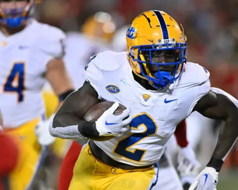 A college football player from the University of Georgia (UGA) in yellow and white uniform carries the football during a game, showcasing athleticism and team spirit, with other players and the stadium background.