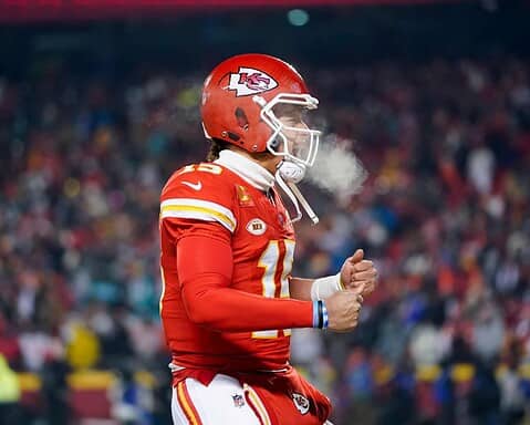 Kansas City Chiefs football player wearing helmet and uniform, celebrating on the field during a game, with fans in the background.