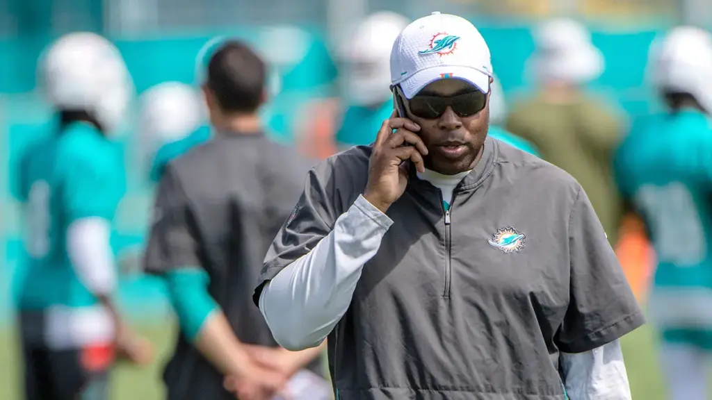 Coach speaking on phone during a practice session with Miami Dolphins staff and players in the background.