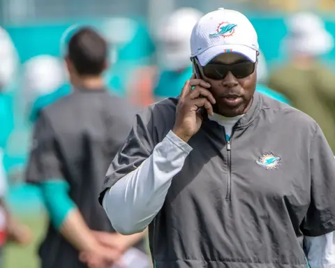 Coach speaking on phone during a practice session with Miami Dolphins staff and players in the background.
