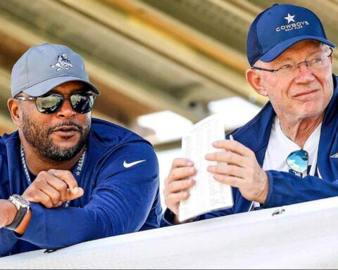 Inside The Star Dallas Cowboys football coaches discussing strategies outdoors in team gear, wearing caps, sunglasses, and jackets with the Dallas Cowboys logo during a game or practice.