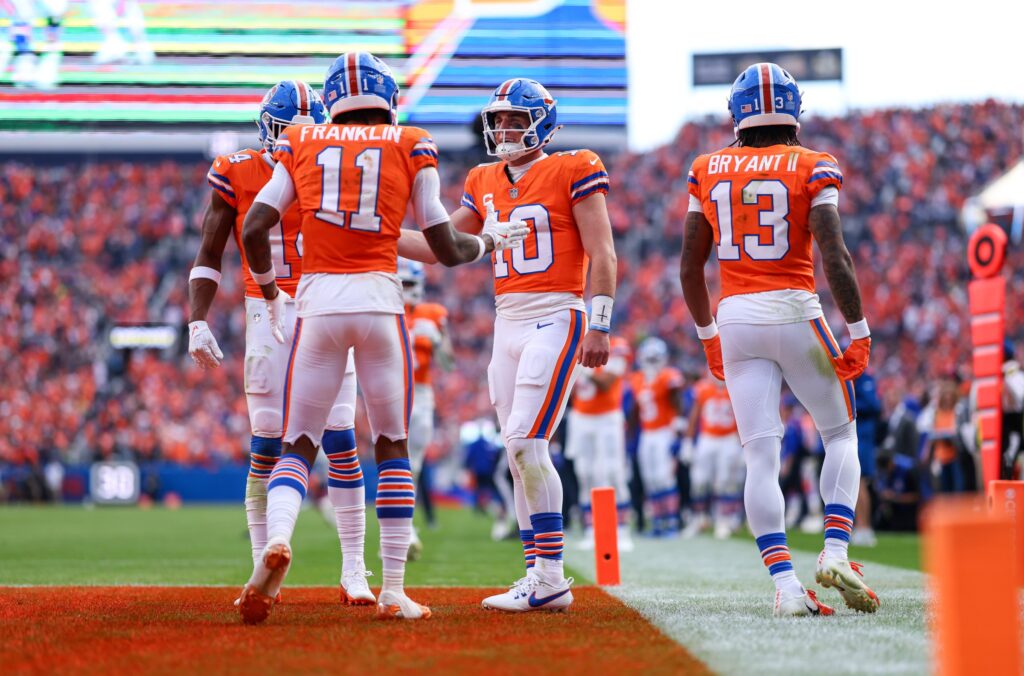 High school football players in orange jerseys celebrating on the field during a game at Empower Field at Mile High, Denver, Colorado. The players are part of the Denver Broncos team, known for their vibrant uniforms.