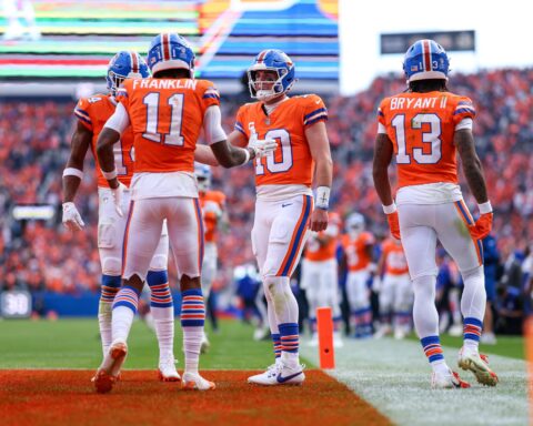 High school football players in orange jerseys celebrating on the field during a game at Empower Field at Mile High, Denver, Colorado. The players are part of the Denver Broncos team, known for their vibrant uniforms.