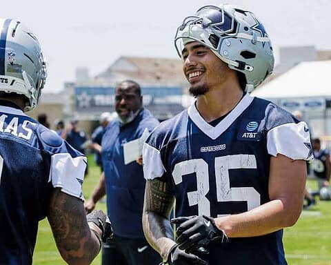 Dallas Cowboys football players during practice, focusing on team strategy and training at the training camp. Players wear helmets and jerseys with the Cowboys logo, engaging in team-building activities outdoors.