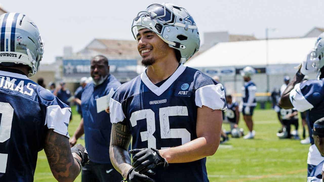 Dallas Cowboys football players during practice, focusing on team strategy and training at the training camp. Players wear helmets and jerseys with the Cowboys logo, engaging in team-building activities outdoors.