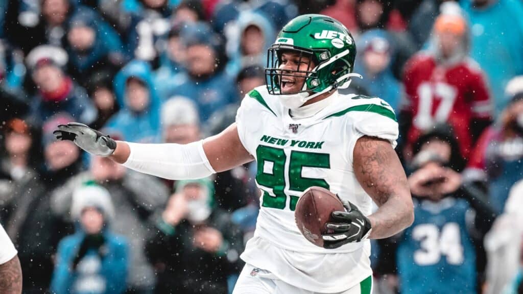 Jets football player holding a football during a game, wearing uniform, helmet, and gloves, with fans in the background in cold weather.
