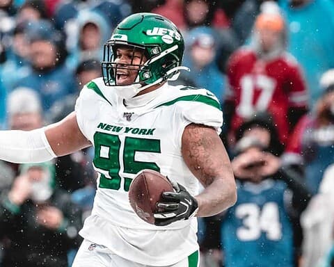 Jets football player holding a football during a game, wearing uniform, helmet, and gloves, with fans in the background in cold weather.