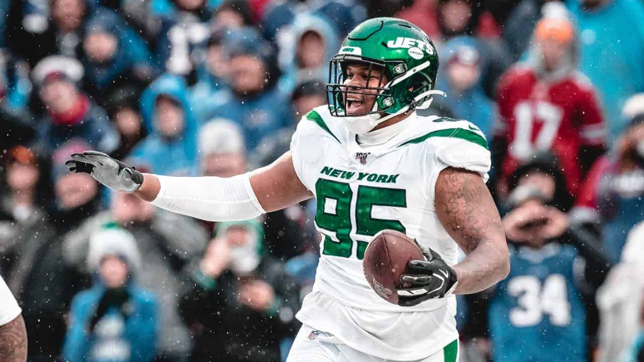 Jets football player holding a football during a game, wearing uniform, helmet, and gloves, with fans in the background in cold weather.